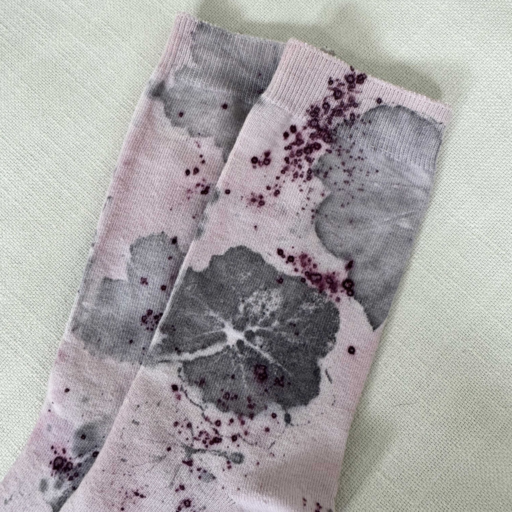 Botanical Geranium Socks - Melo Handmades. Pair of lavender-toned socks with botanical dye patterns, worn on a bed with soft white linen. 